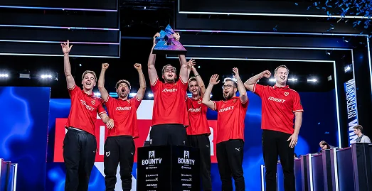 Een esports-team viert op het podium een kampioenschapsoverwinning met een trofee en opgeheven armen voor een groot publiek.